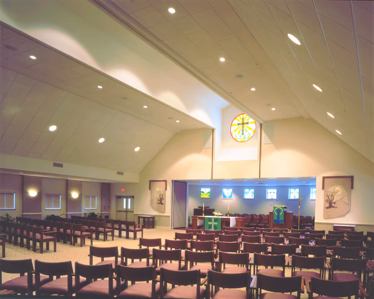 Interior view of a chapel or worship area with rows of wooden chairs facing an altar. The altar is decorated with green cloths featuring religious symbols, and there are stained glass windows above and behind it, including a circular one with a cross. The ceiling is high with recessed lighting, and there are wall hangings on either side of the altar.