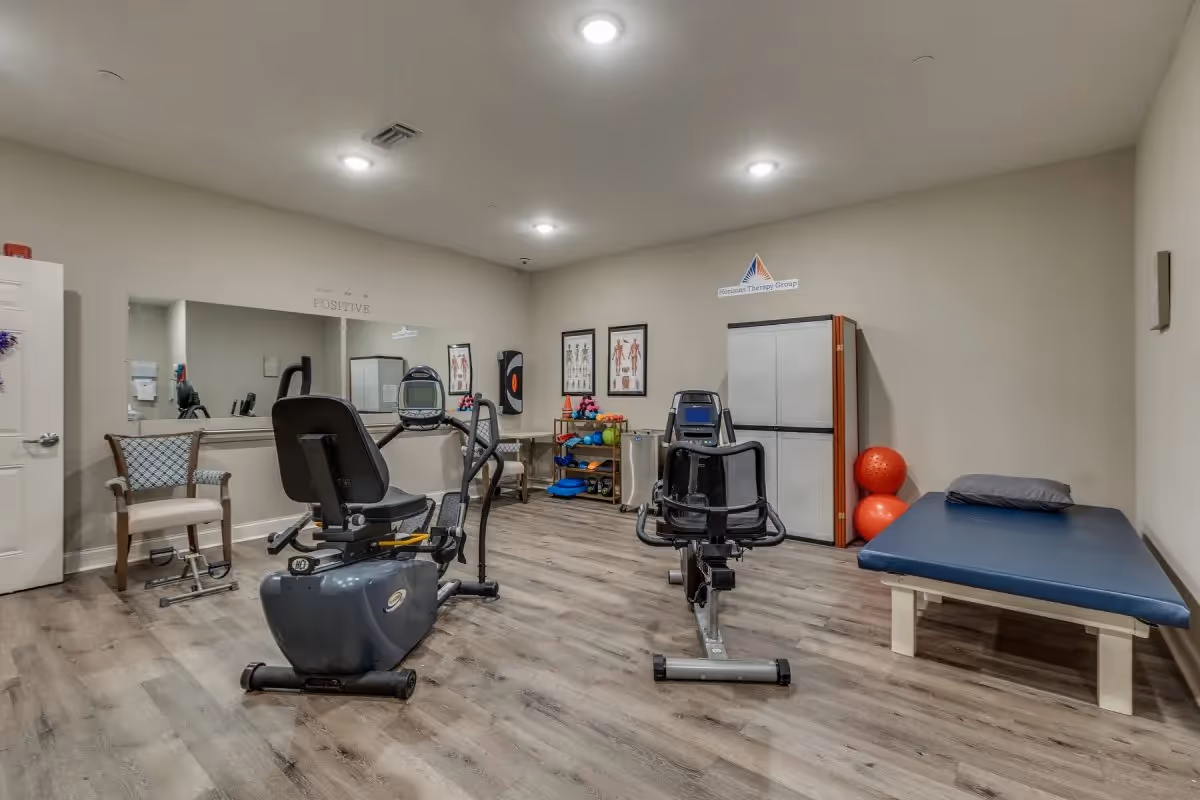 A therapy or exercise room in an assisted living facility with two stationary exercise bikes, a padded therapy table with a pillow, a chair, a large mirror on the wall, anatomical charts, and shelves holding various exercise equipment including colorful dumbbells and exercise balls.