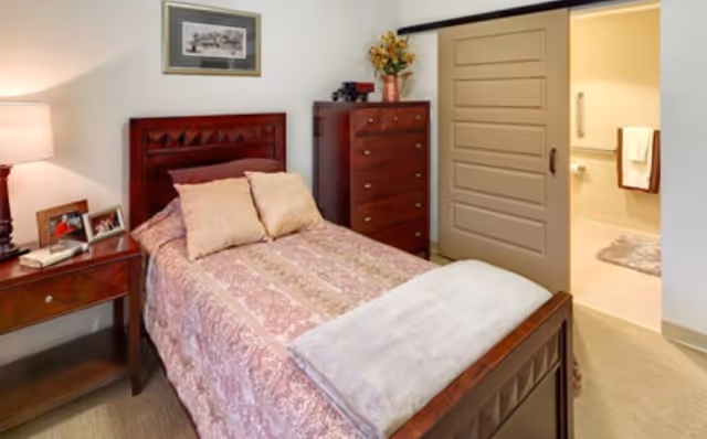 Single bed with a patterned bedspread in a furnished bedroom with a nightstand, dresser, lamp, and an open sliding door to a bathroom.