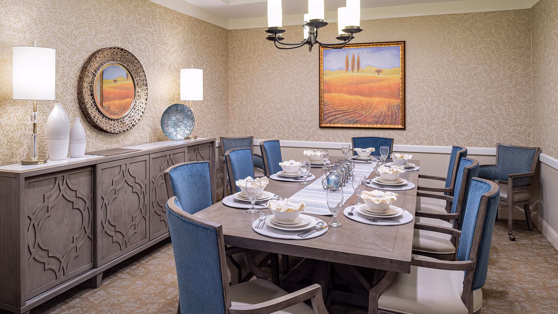 A dining room with a rectangular wooden table set for eight people. The table is decorated with white plates, bowls, napkins, and glassware. Surrounding the table are blue upholstered chairs with wooden frames. Against the wall is a wooden sideboard with decorative vases and a round mirror above it. The walls have a subtle patterned wallpaper, and a framed landscape painting hangs on the far wall. A chandelier with five lights hangs above the table.