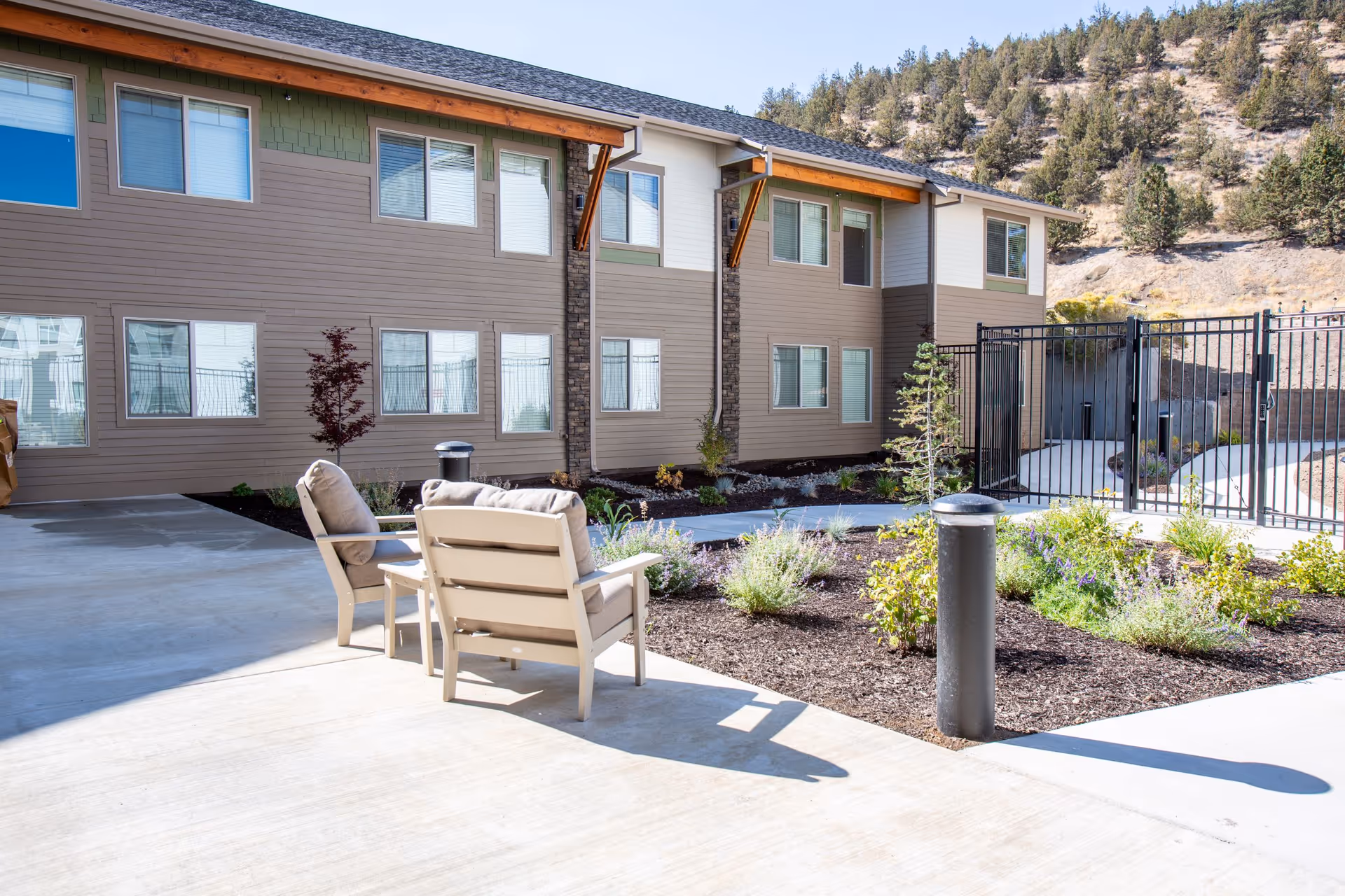 Outdoor patio area at Cascades of Bend Retirement Community with cushioned chairs and a small table on a concrete surface, surrounded by landscaped garden beds and a two-story building in the background under a clear sky.