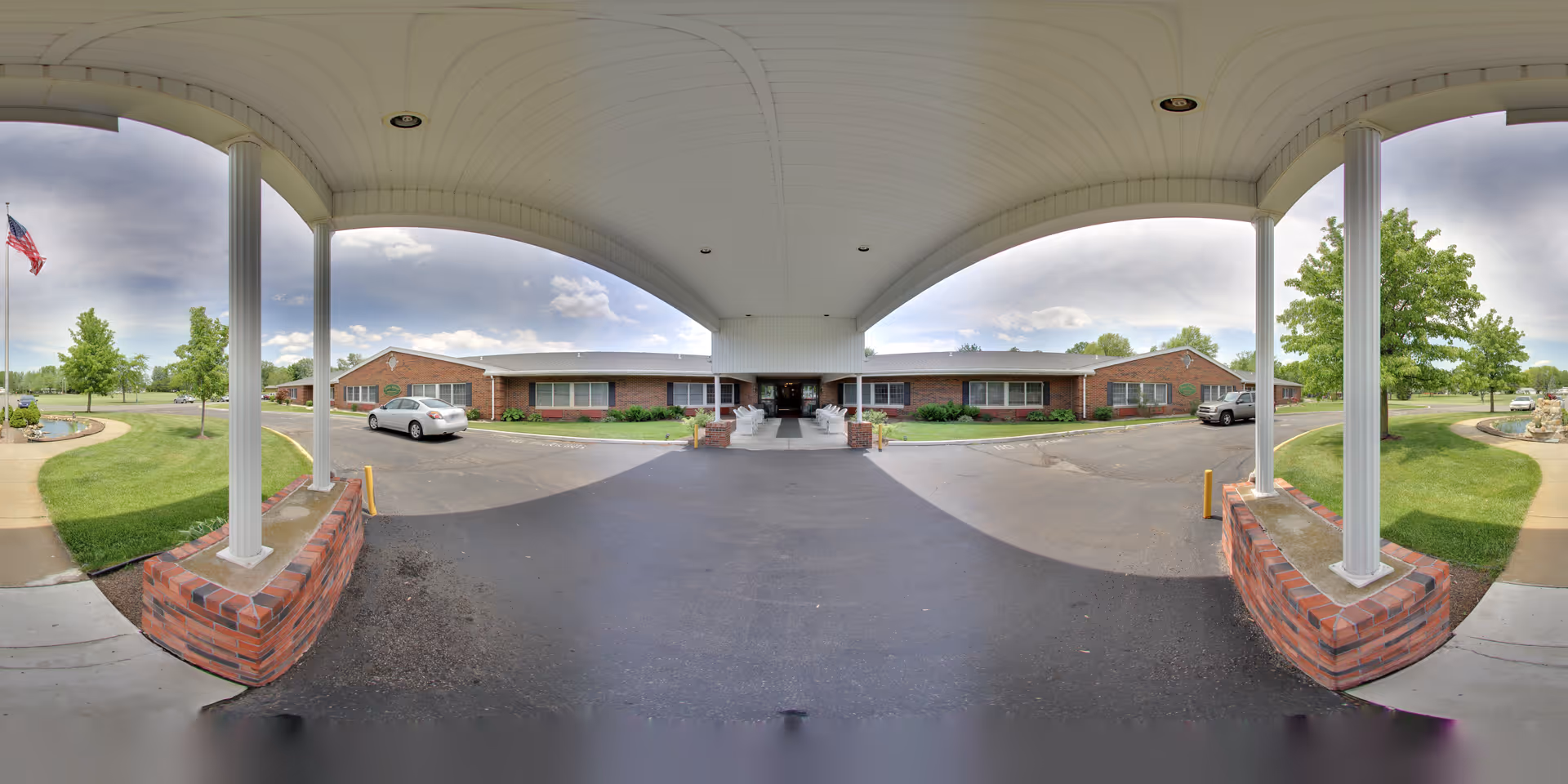 Covered driveway entrance to a single-story brick building with white columns supporting the roof. There are parked cars on either side of the driveway, green lawns with trees and a flagpole with an American flag on the left side. The sky is partly cloudy.