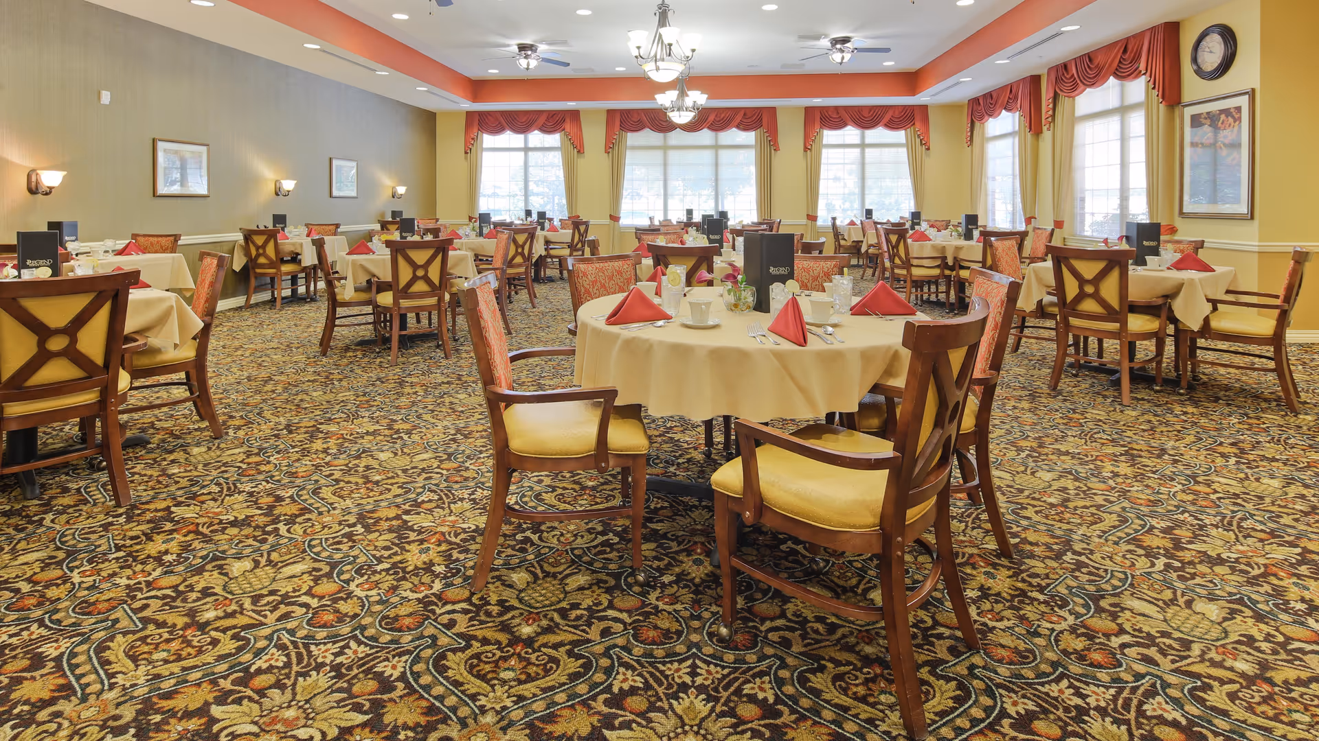 A spacious dining room with multiple round and square tables covered with beige tablecloths. Each table is set with red folded napkins, cups, and silverware. The room has large windows with red valances, patterned carpet, and warm yellow walls. Wooden chairs with yellow cushions surround the tables.