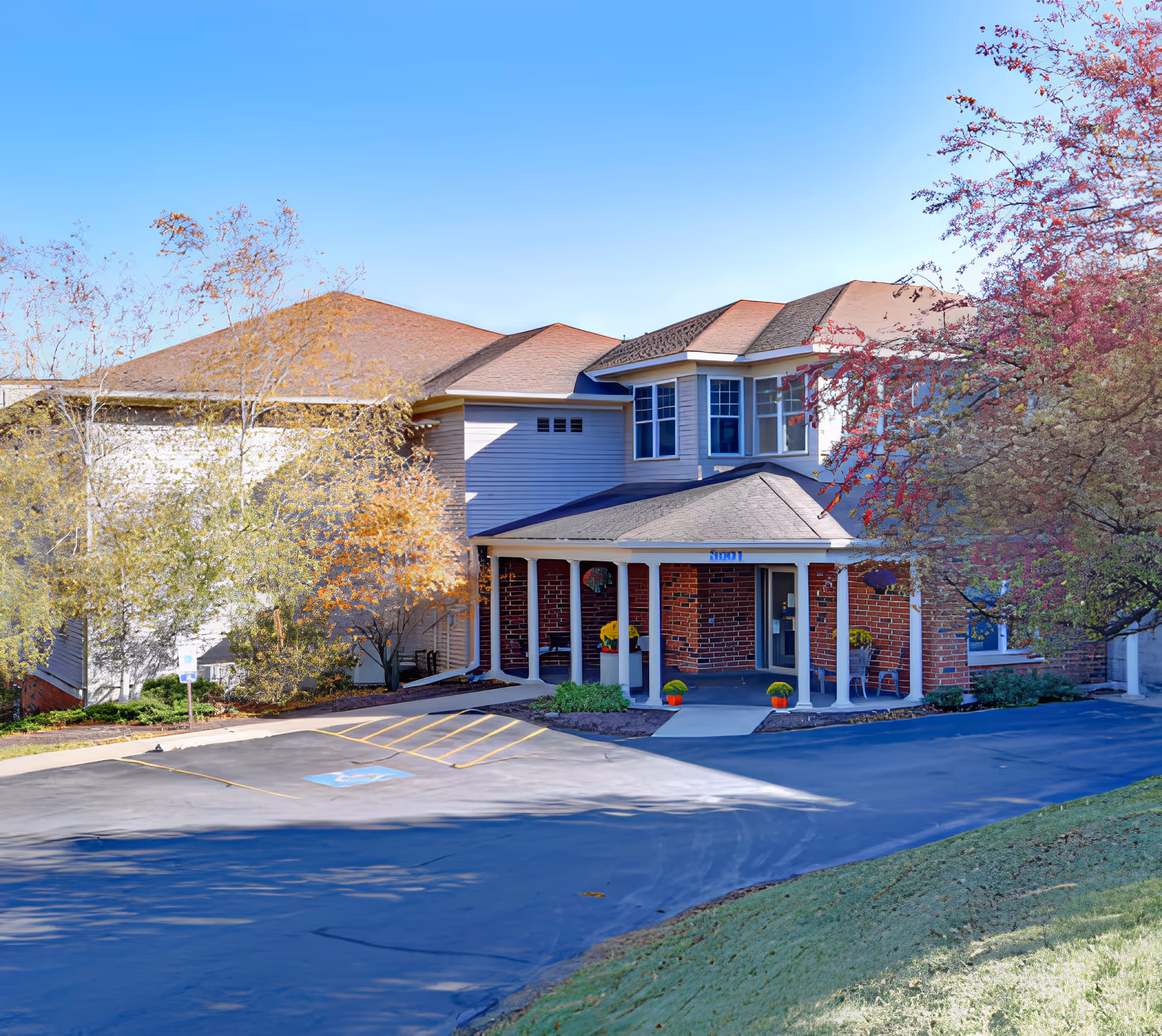 Exterior view of a senior living facility named Applegate Terrace, showing a two-story building with a covered entrance supported by white columns. The building has a combination of brick and siding exterior, surrounded by trees with autumn foliage and a clear blue sky overhead. There is a parking area with a designated handicapped parking space in front of the entrance.