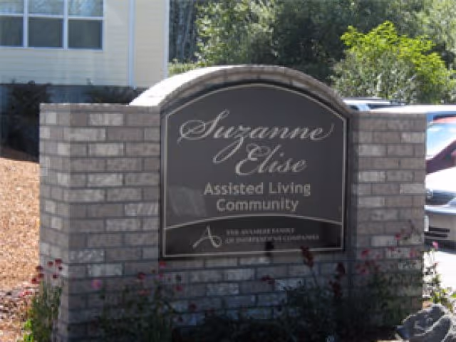 Stone entrance sign reading "Suzanne Elise Assisted Living Community" set in brick with landscaping and parked cars behind it.