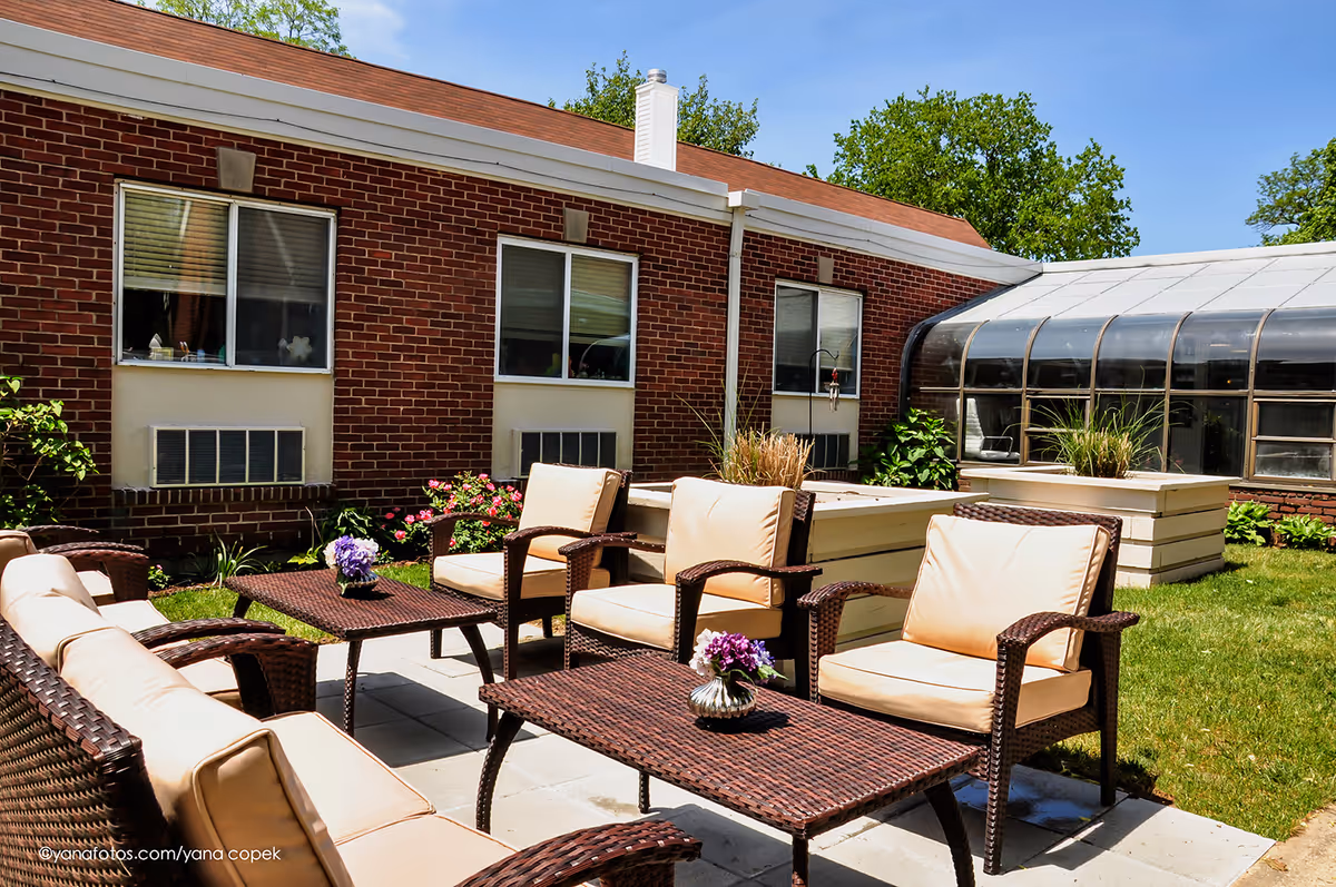 Outdoor patio area with cushioned wicker chairs and tables arranged on a stone-paved surface next to a brick building with windows. There are flower pots on the tables and greenery around the patio under a clear blue sky.