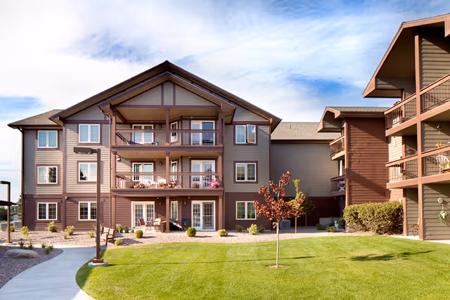 Front exterior of a three-story senior living building with balconies, landscaped lawn, and walkways.