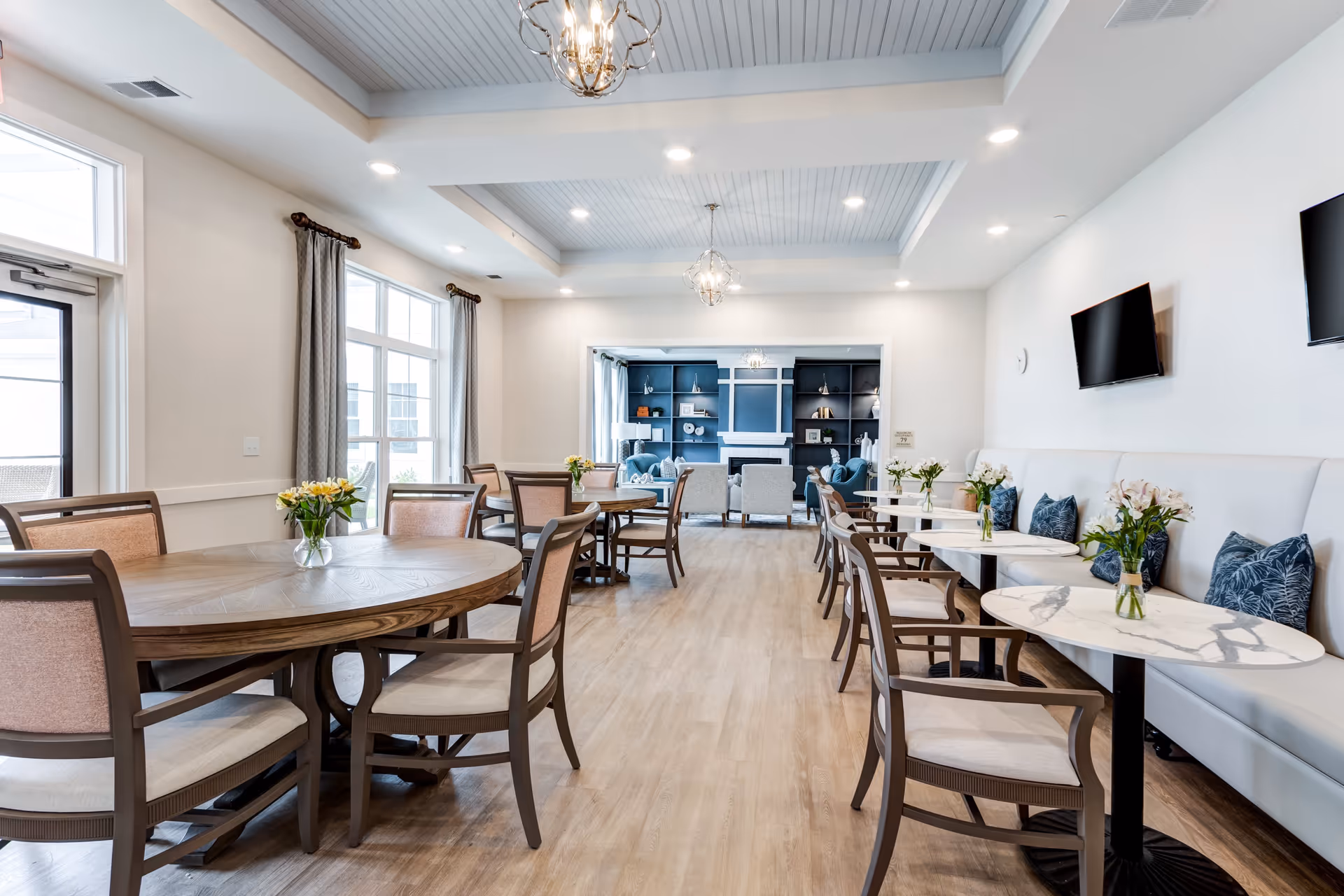A bright and spacious dining area with round wooden tables and chairs arranged neatly. Each table has a small vase with flowers. The room has large windows with curtains allowing natural light to fill the space. In the background, there is a cozy seating area with armchairs and a fireplace surrounded by built-in shelves. The ceiling features recessed lighting and decorative light fixtures.