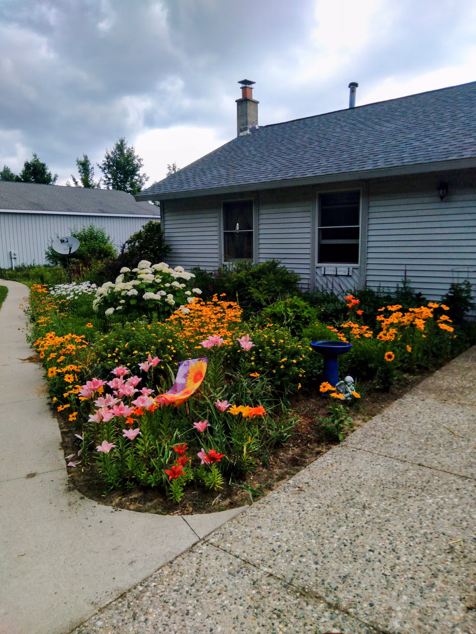 Front exterior of a single-story house with a colorful flowerbed alongside a concrete walkway under a cloudy sky.