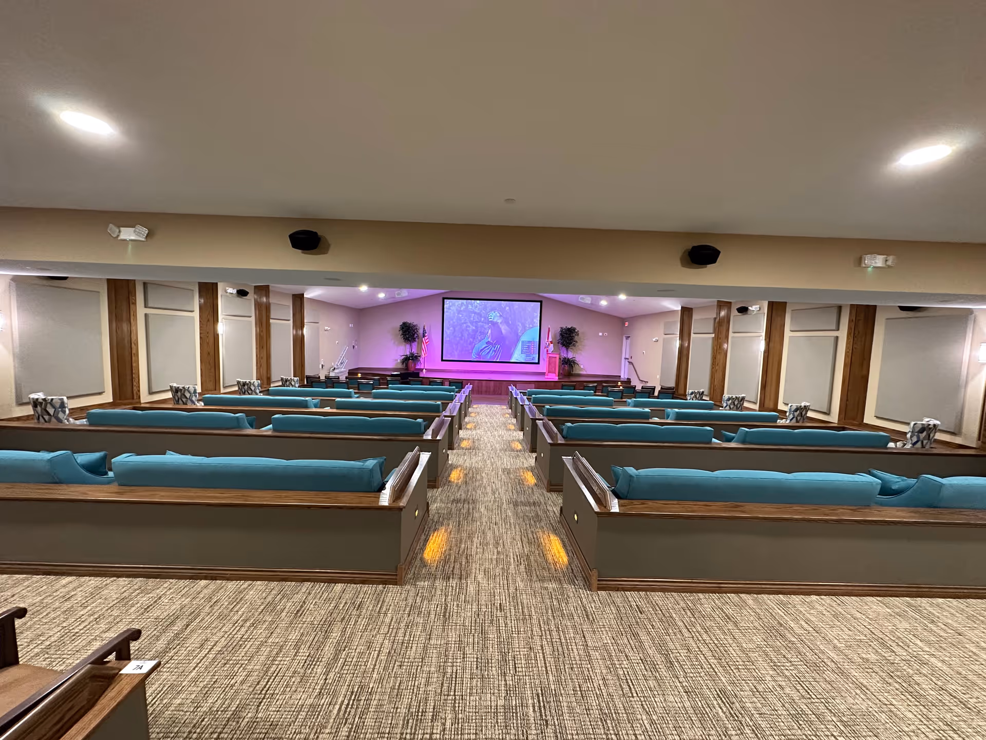 Interior view of a large room with multiple rows of wooden pew-style seating with teal cushions facing a stage with a large screen displaying an image. The room has beige walls with sound panels and recessed lighting in the ceiling.