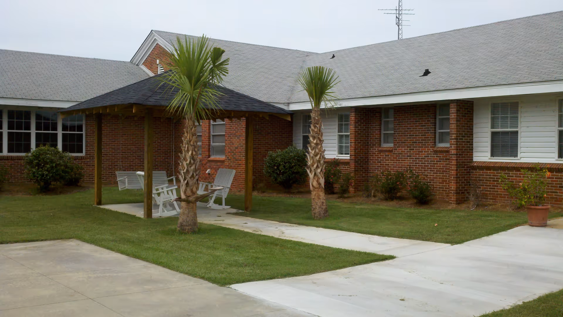 Brick retirement community courtyard with a covered seating area, two palm trees, white chairs, and concrete walkways.
