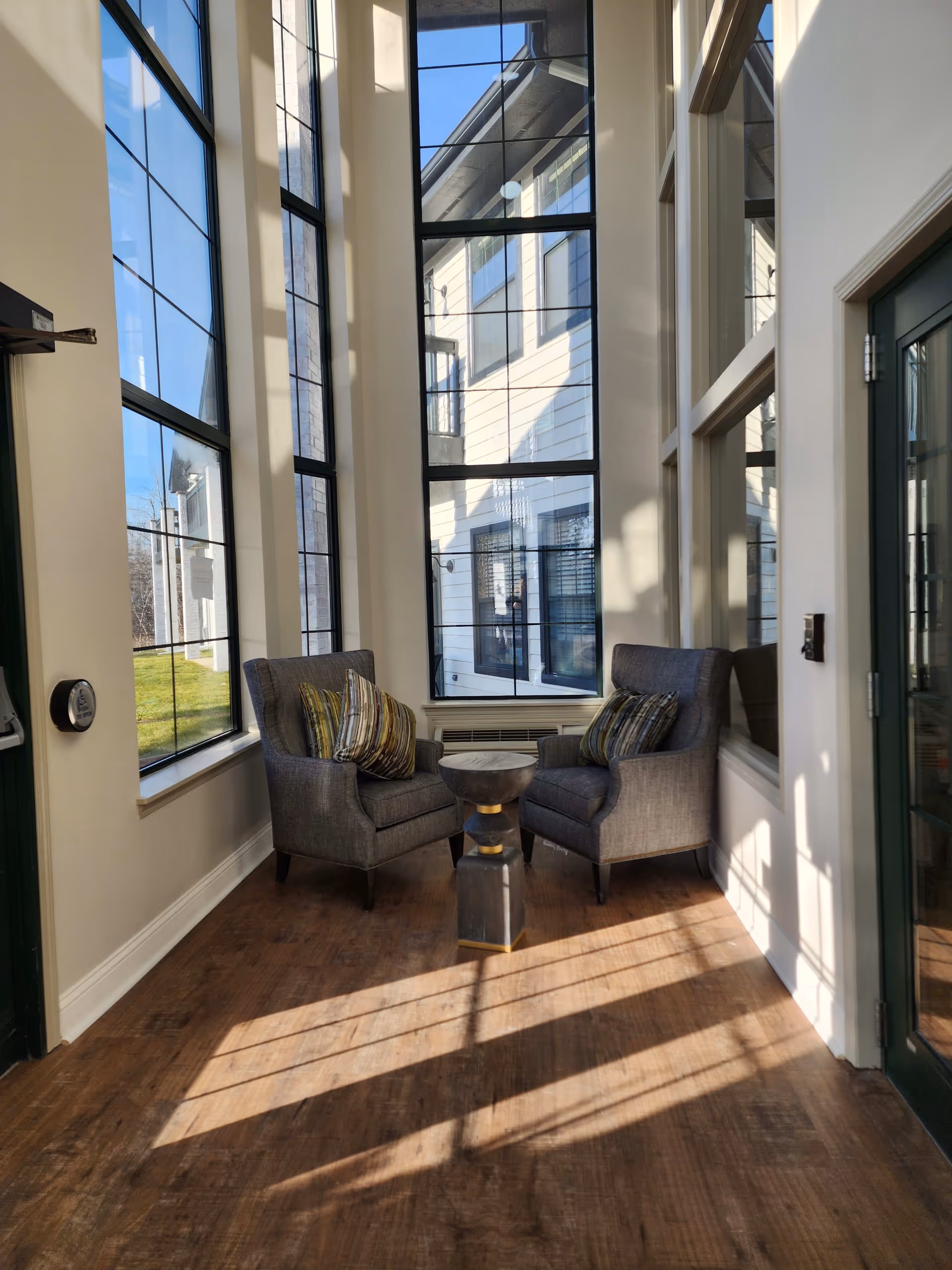 A sunlit lobby seating area featuring two gray armchairs with patterned cushions and a small table, surrounded by large windows that allow natural light to fill the space.