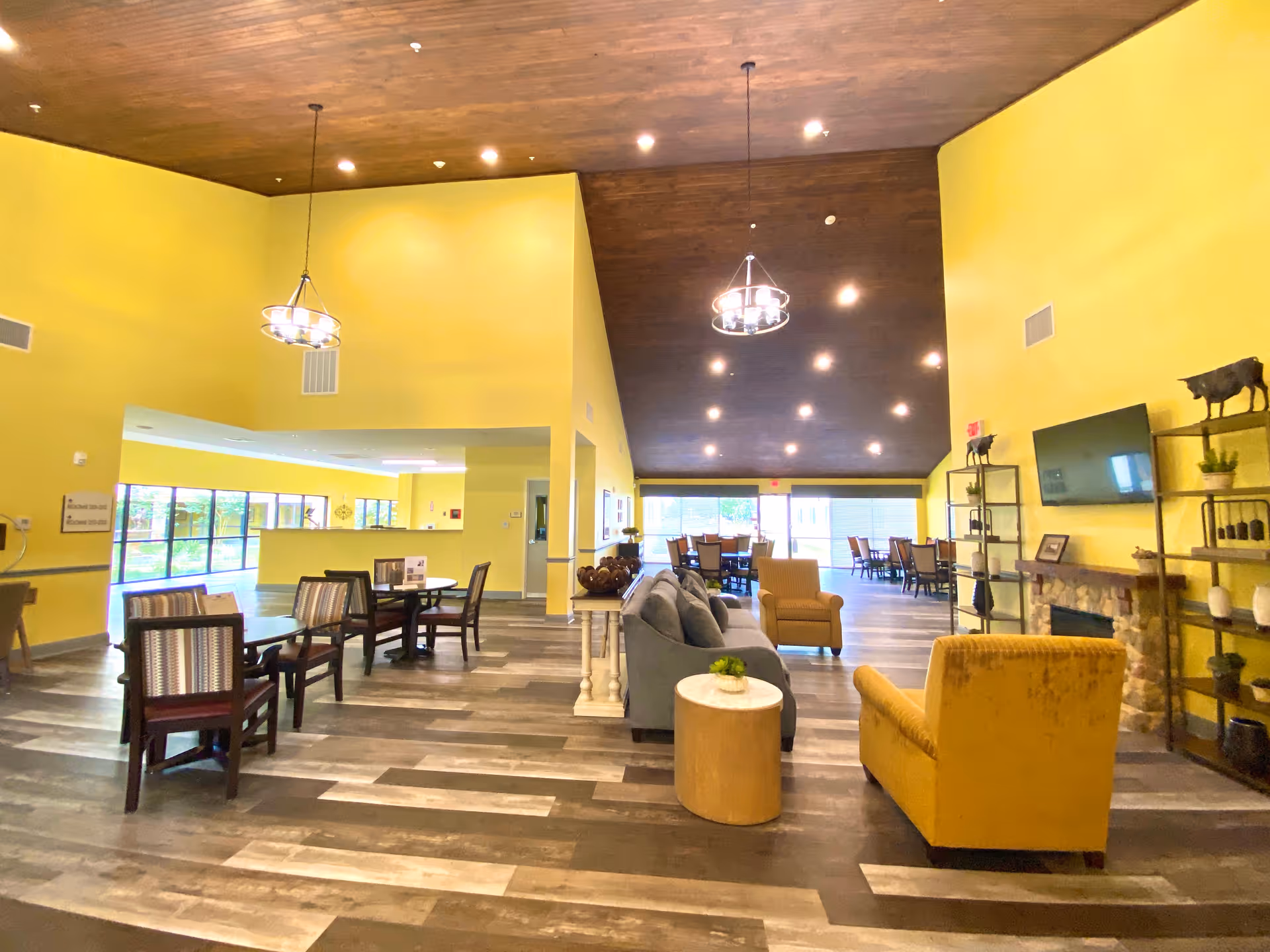 A spacious senior living facility common area with yellow walls and a wooden ceiling. The room features multiple seating arrangements including a gray sofa, yellow armchairs, and several tables with chairs. There is a mounted flat-screen TV above a stone fireplace, decorative shelves with plants and ornaments, and large windows letting in natural light.