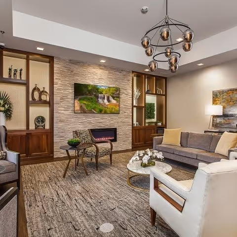 A modern living room with a gray sofa, white armchair, and a patterned chair around a round coffee table with white flowers. The room features a textured stone wall with a mounted flat-screen TV above a built-in electric fireplace. Wooden built-in shelves with decorative items flank the fireplace. A contemporary chandelier hangs from the ceiling, and a table lamp and abstract painting are visible on the right side.