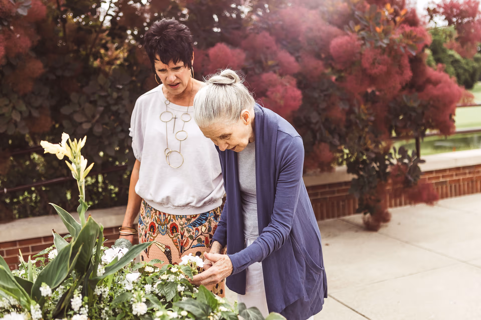 Two women outdoors near a garden bed with various green plants and white flowers. One woman with gray hair tied back is closely examining the flowers, while the other woman with short dark hair stands beside her, looking at the plants. Behind them are bushes with reddish foliage and a brick wall.
