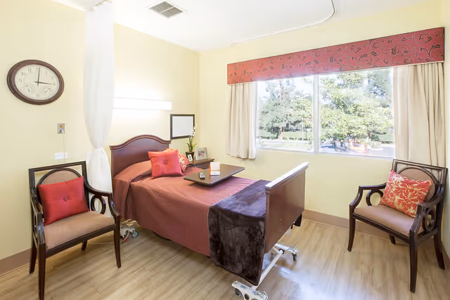 A bright and clean senior living bedroom with a single hospital-style bed covered in a red bedspread and a dark brown blanket at the foot. There are two wooden chairs with red cushions on either side of the bed. A large window with beige curtains and a red valance lets in natural light, showing greenery outside. A wall clock and a small mirror are mounted on the pale yellow walls.