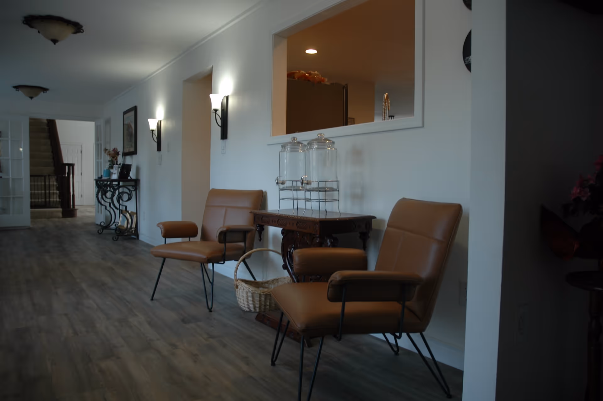 A hallway with two brown leather chairs and a small ornate wooden table between them holding two glass beverage dispensers. The hallway has wooden flooring, white walls with two wall sconces, and a window opening into another room. In the background, there is a staircase and a console table with decorative items.