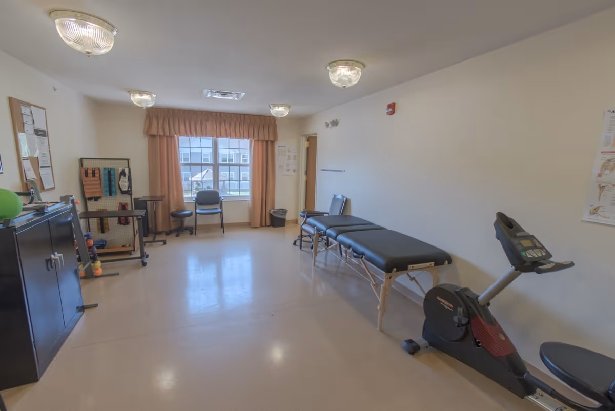 Bright therapy/fitness room with an exercise bike, two treatment tables, chairs and small exercise equipment near a window.