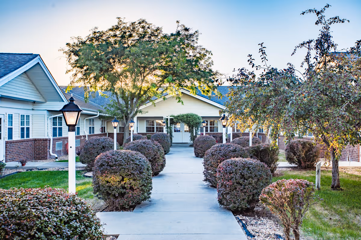 A paved walkway lined with trimmed bushes and lamp posts leads to the entrance of a single-story building with white siding and brick accents. Trees flank the walkway, and sunlight filters through the leaves, creating a serene outdoor setting.