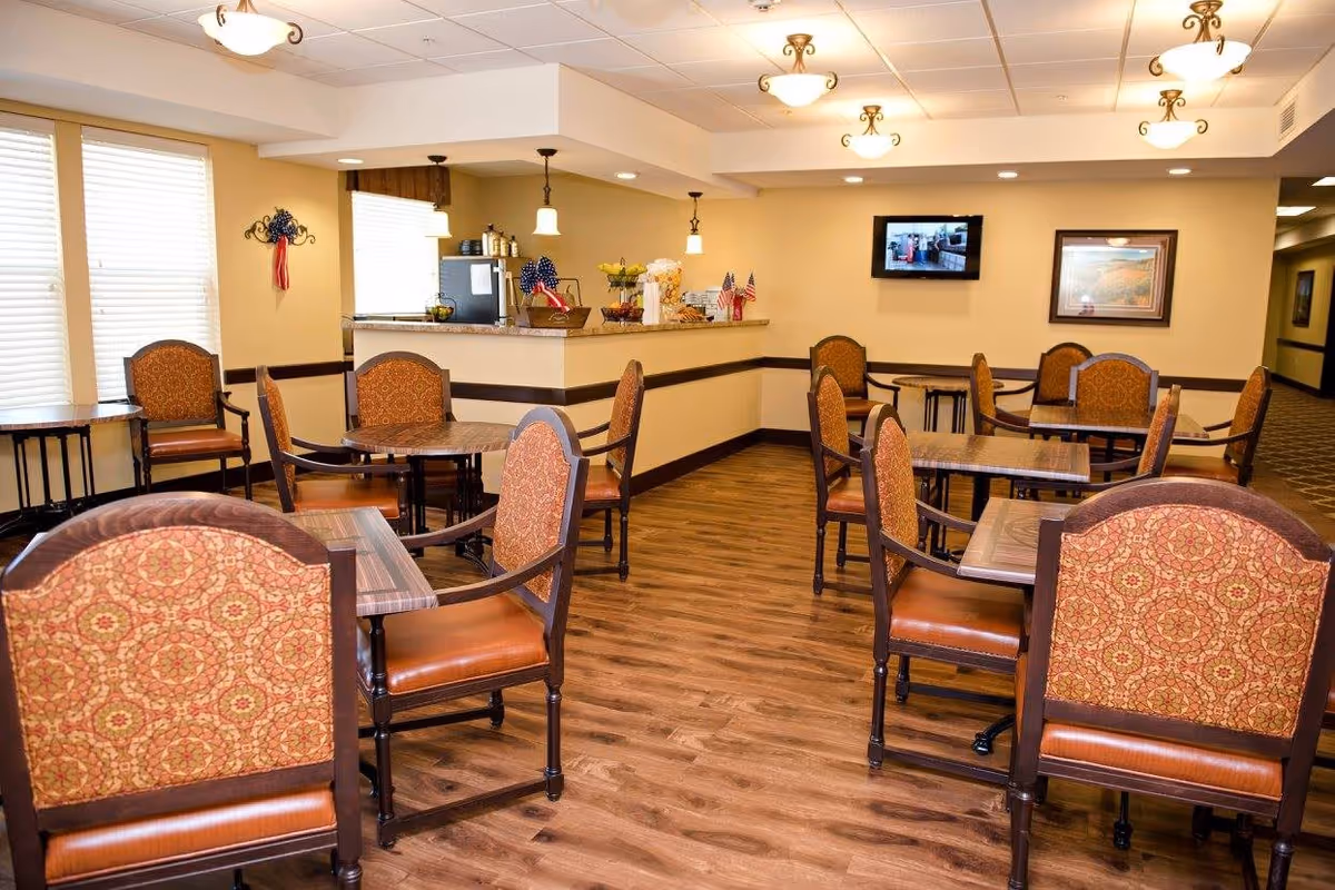 A dining area in a senior living facility with several wooden tables and chairs upholstered in patterned fabric. The room has wood flooring, beige walls, and ceiling lights. There is a counter with small American flags and baskets, a wall-mounted TV, and framed artwork on the walls.