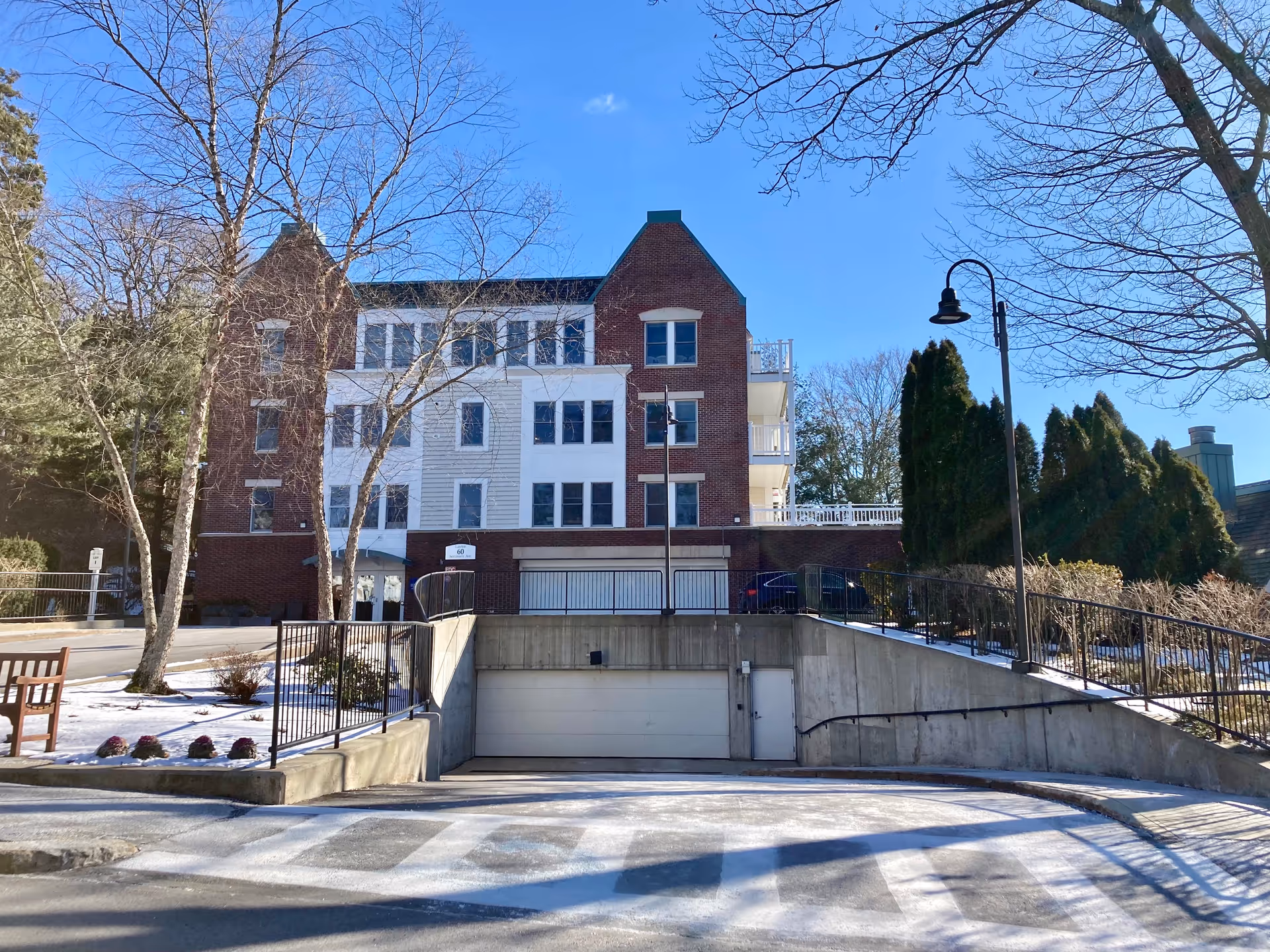 Exterior view of a multi-story brick and white-paneled building with several windows, a garage entrance at the lower level, leafless trees, and a clear blue sky.