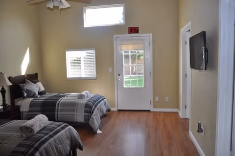 A bright bedroom with two single beds covered in gray and white plaid bedding, each with a rolled towel on top. The room has light yellow walls, a wooden floor, a ceiling fan with lights, a window with white blinds, and a glass door with an exit sign above it. A flat-screen TV is mounted on the wall to the right.