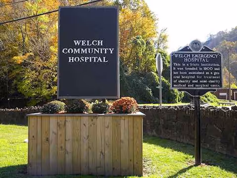 Outdoor scene with Welch Community Hospital signs, a wooden planter, and surrounding trees.