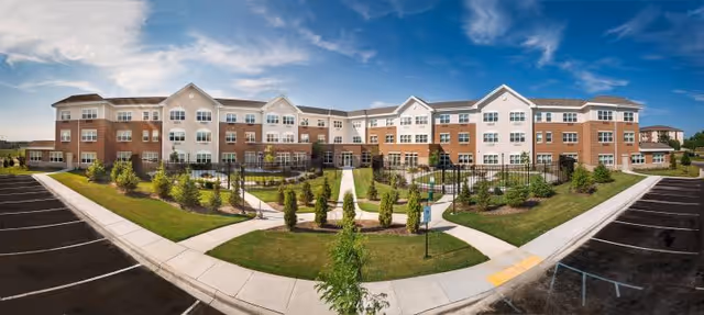 Wide exterior view of a three-story brick-and-siding retirement community with a landscaped courtyard and surrounding parking.