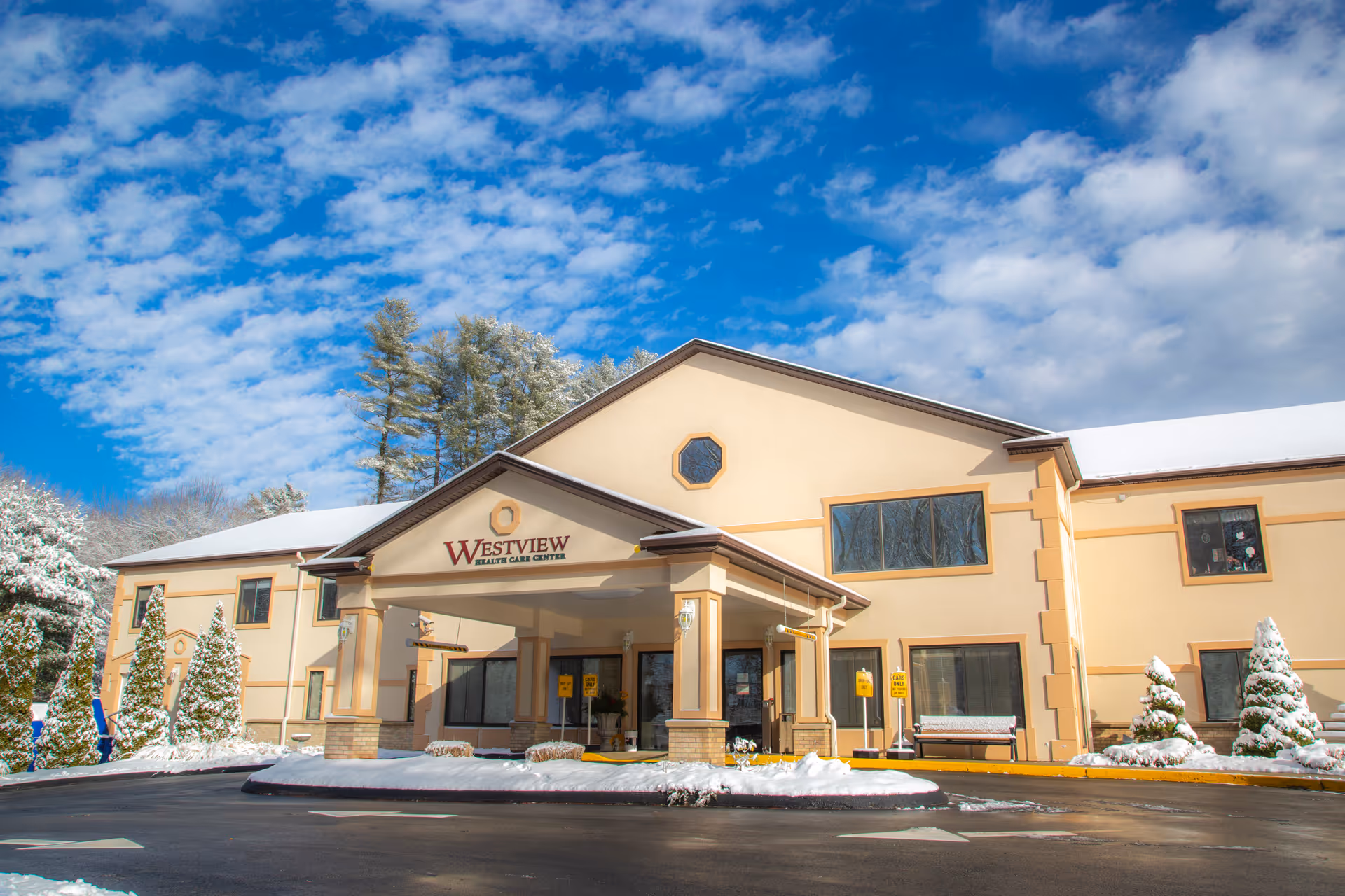 Exterior front view of Westview Health Care Center building on a snowy day with a clear blue sky and some clouds. The building is beige with brown trim and has a covered entrance with columns. Snow-covered trees and shrubs are visible around the building.