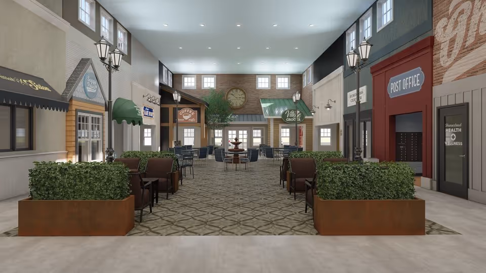 Indoor common area designed to resemble a small village street with storefronts including a post office, coffee cafe, village grocer, and health and wellness center. The space features seating areas with chairs and tables, decorative street lamps, potted plants, and a large clock on a brick wall at the far end.