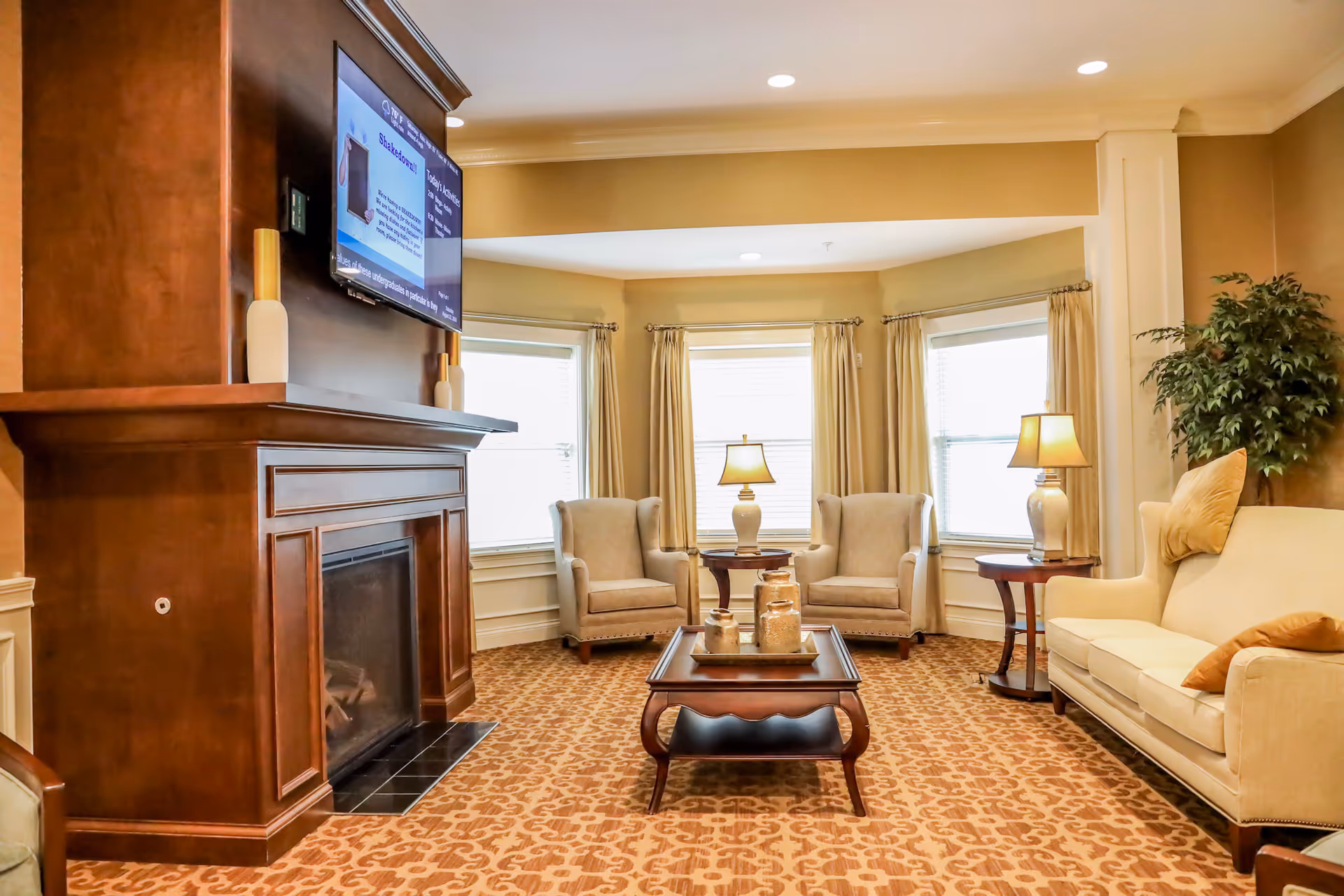 A cozy living room area with a large wooden fireplace on the left, a wall-mounted TV above it, two beige armchairs and a small round table with a lamp in front of three windows with curtains. On the right, there is a beige sofa with cushions, a side table with a lamp, and a tall green plant in the corner. A wooden coffee table with decorative jars is centered on a patterned carpet.