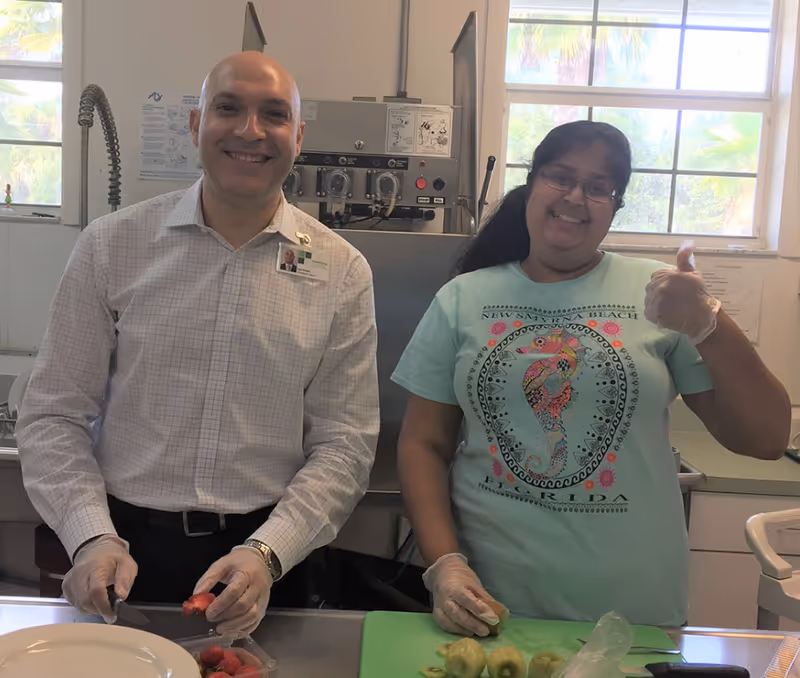 Two smiling staff members wearing gloves prepare fruit on a kitchen counter inside an assisted living facility.