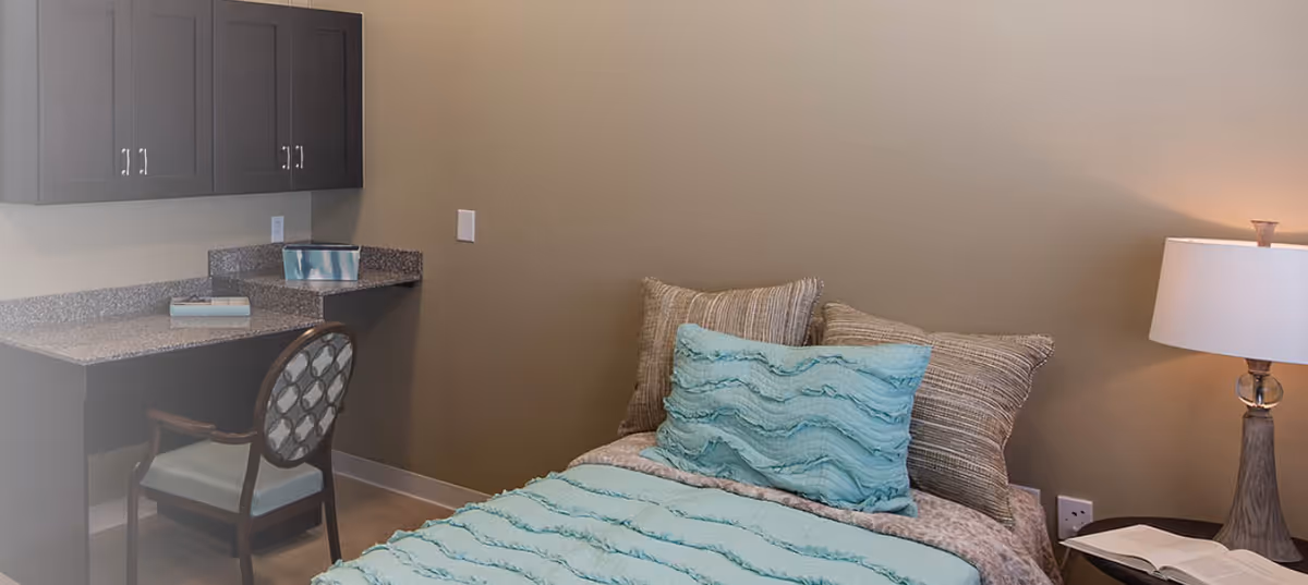 Small bedroom with a teal-covered bed, decorative pillows, a bedside lamp, and a built-in desk with cabinets and a chair.