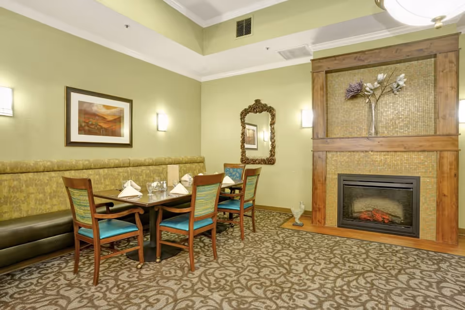 Dining area with a set table, banquette seating and chairs in front of a decorative fireplace and mirror.