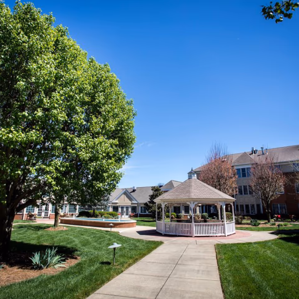A well-maintained outdoor courtyard at Village Manor featuring a white gazebo, a paved walkway, green grass, trees with fresh leaves, and a clear blue sky.