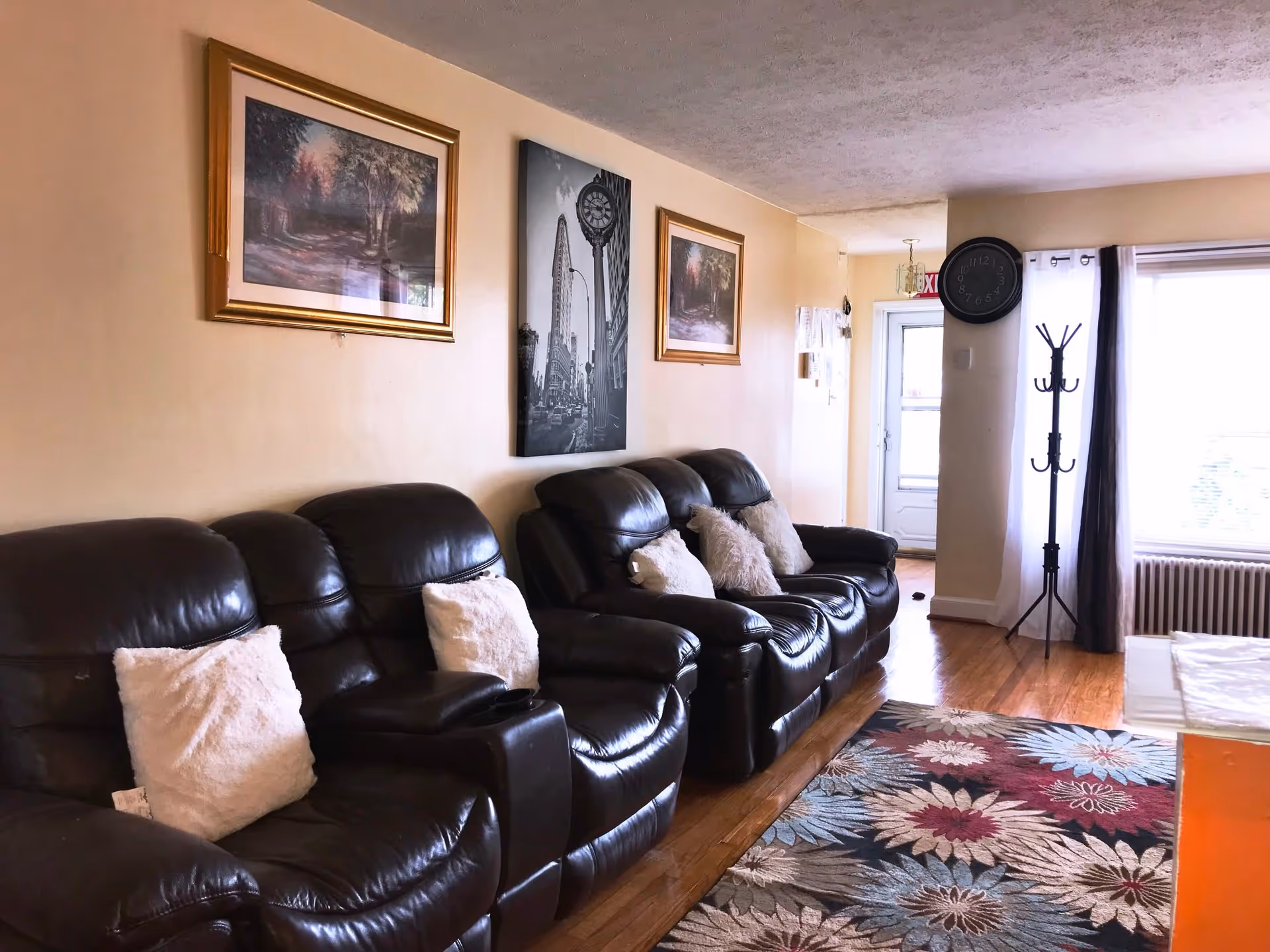 Bright living room with a row of dark leather recliners, decorative pillows, framed wall art, a coat rack, and a floral area rug.
