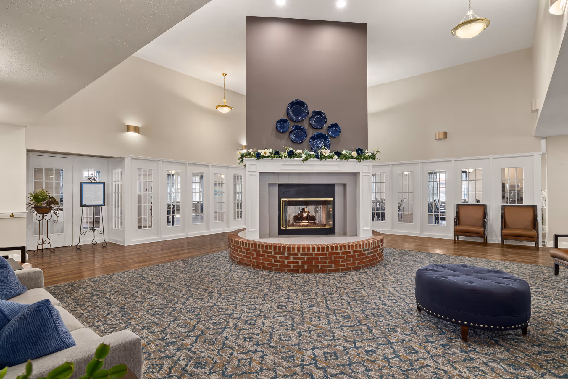 Spacious senior living facility common area with a large central fireplace featuring a brick base and decorated mantle. The room has high vaulted ceilings, a patterned carpet, and seating including a gray sofa with blue pillows and a blue ottoman. Glass-paneled doors line the walls, allowing natural light to enter the space.