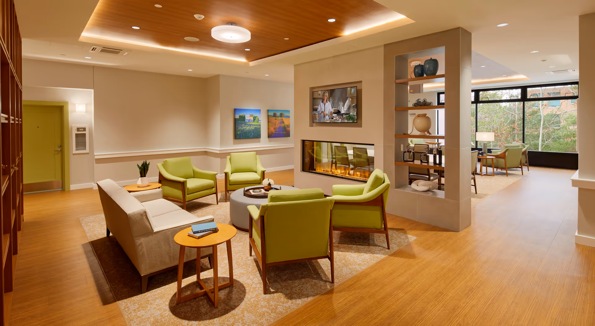 A modern and cozy living room area with a beige sofa, four green armchairs arranged around a circular ottoman, and a small wooden side table with books. The room features a built-in fireplace with a TV mounted above it, decorative shelves with vases, and large windows letting in natural light. The flooring is wooden, and the ceiling has recessed lighting with a wooden panel section.