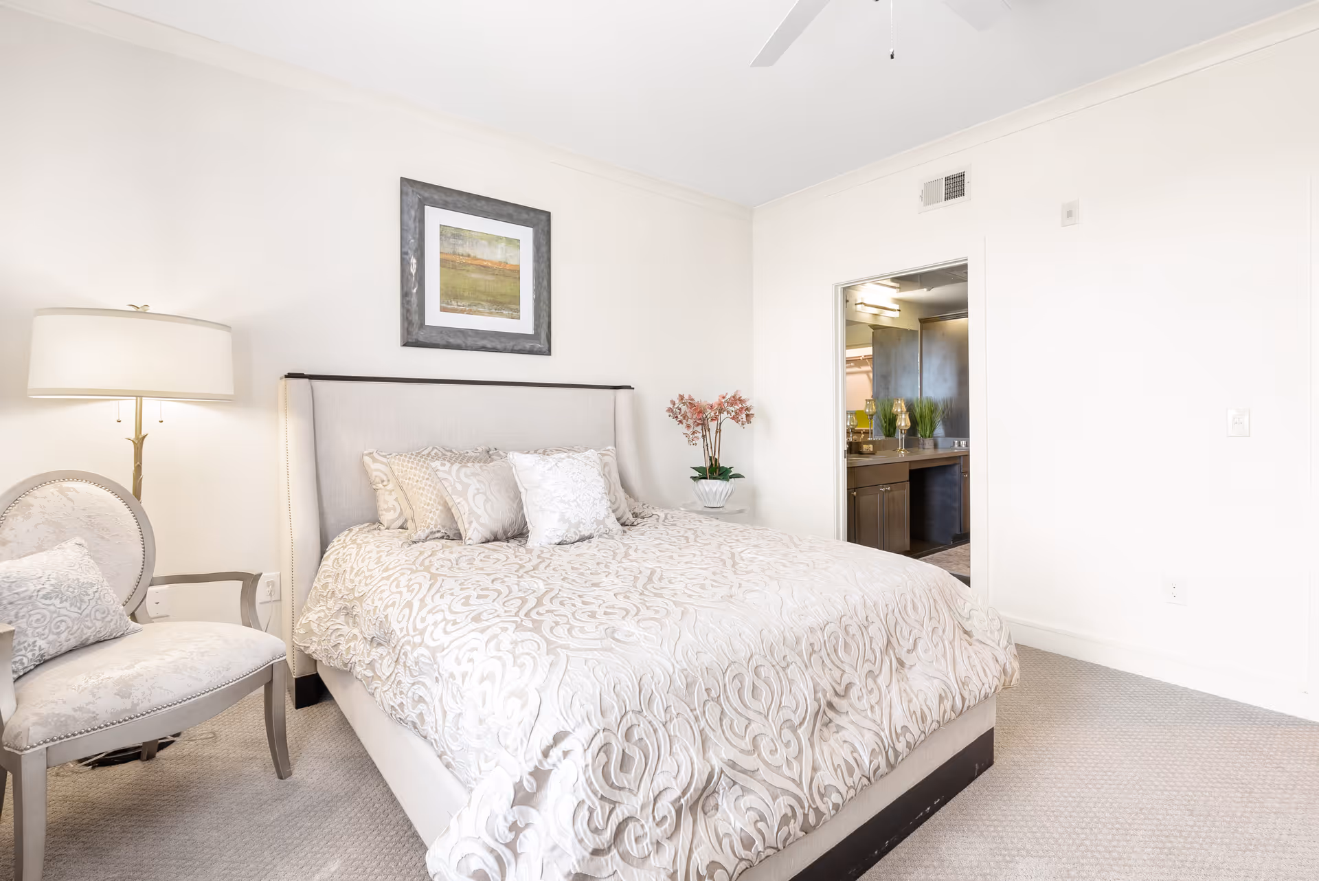 A bright bedroom with a large bed covered in a patterned beige and white comforter and multiple pillows. To the left of the bed is a cushioned chair with a decorative pillow and a tall floor lamp. A framed landscape painting hangs on the wall above the bed. An open doorway reveals a glimpse of a bathroom with a sink and mirror. The room has light-colored walls and carpeted flooring.
