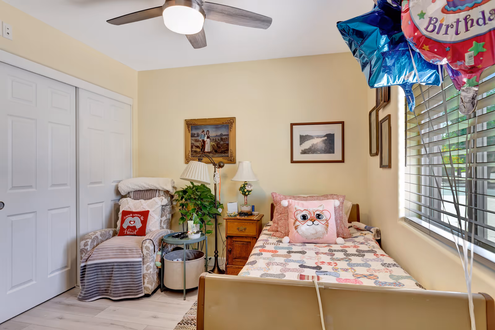 A cozy bedroom in an assisted senior living facility featuring a single bed with a colorful patchwork quilt and a decorative cat pillow. Next to the bed is a wooden nightstand with two lamps and some small items. A comfortable armchair with a striped blanket and a red pillow that says 'Friends warm the heart' is positioned near a closet with white sliding doors. There are framed pictures on the light yellow walls and a ceiling fan with a light fixture. A window with blinds lets in natural light, and several colorful birthday balloons are tied near the window.