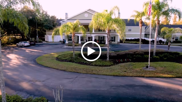 Front exterior view of a senior living facility named Madison at Oviedo, featuring a circular driveway with palm trees and an American flag in front of the building.