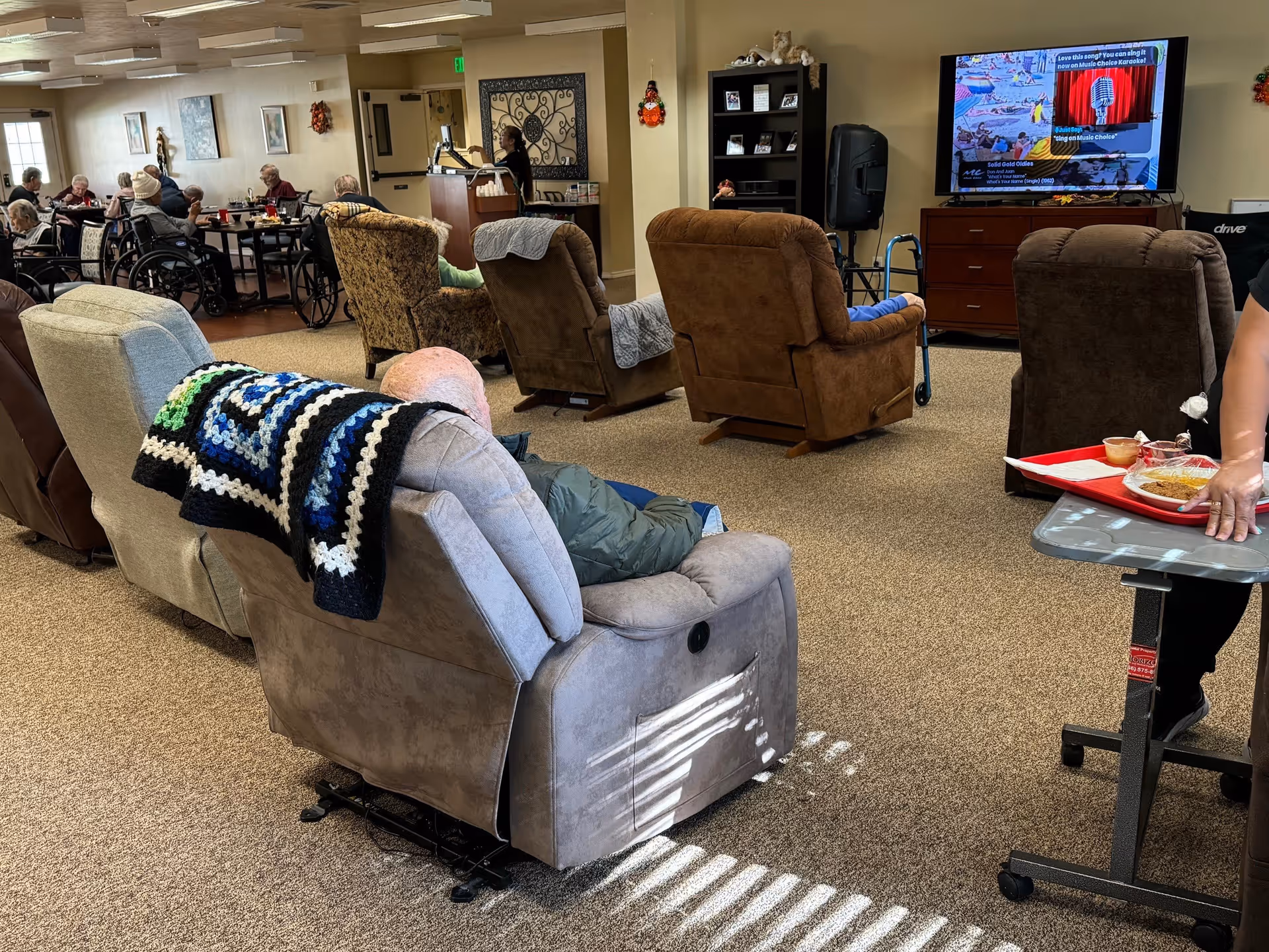 A common area in a senior living facility with several elderly residents seated in recliners and wheelchairs. Some residents are watching a large TV screen mounted on the wall, while others are seated at tables in the background. A staff member is partially visible on the right side, holding a tray with food. The room has beige walls, carpeted floors, and various pieces of furniture including recliners, tables, and a cabinet with decorations.