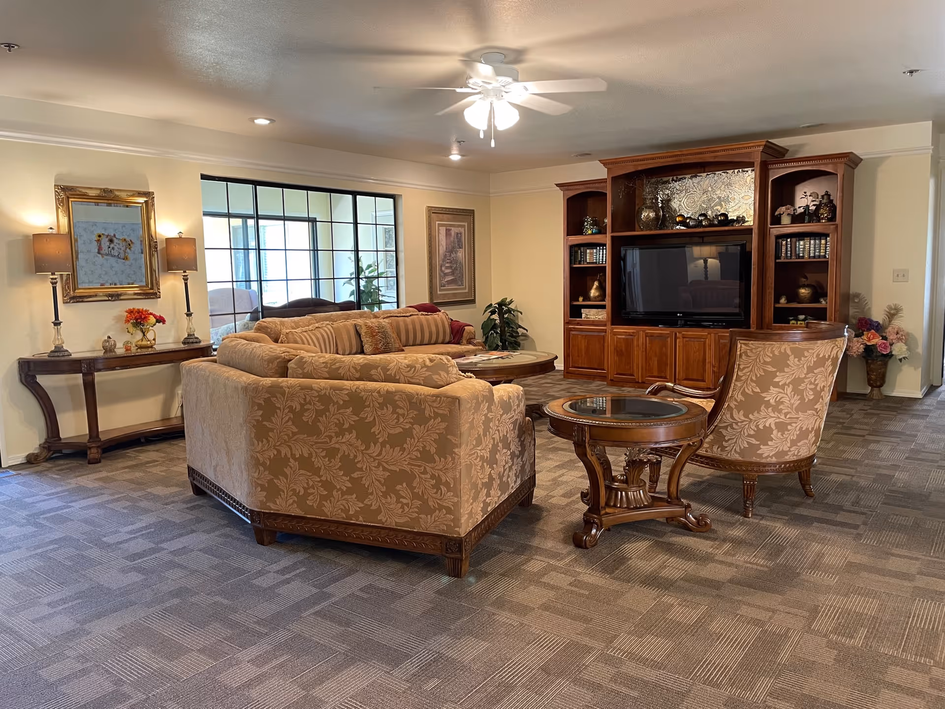 A cozy living room with beige patterned sofas and armchairs arranged around a wooden coffee table. There is a large wooden entertainment center with a flat-screen TV and decorative items. The room has carpeted flooring, a ceiling fan with lights, and a console table with two lamps and a framed mirror on the wall.