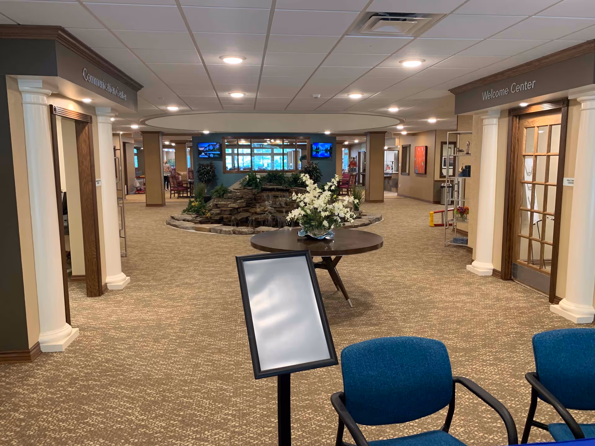 Interior view of a senior living facility lobby area with a round table holding a floral arrangement in the center. There is a small rock water feature behind the table, two blue chairs in the foreground, and signs indicating a Welcome Center on the right and a Community Activity Center on the left. The space is well-lit with ceiling lights and has carpeted floors.