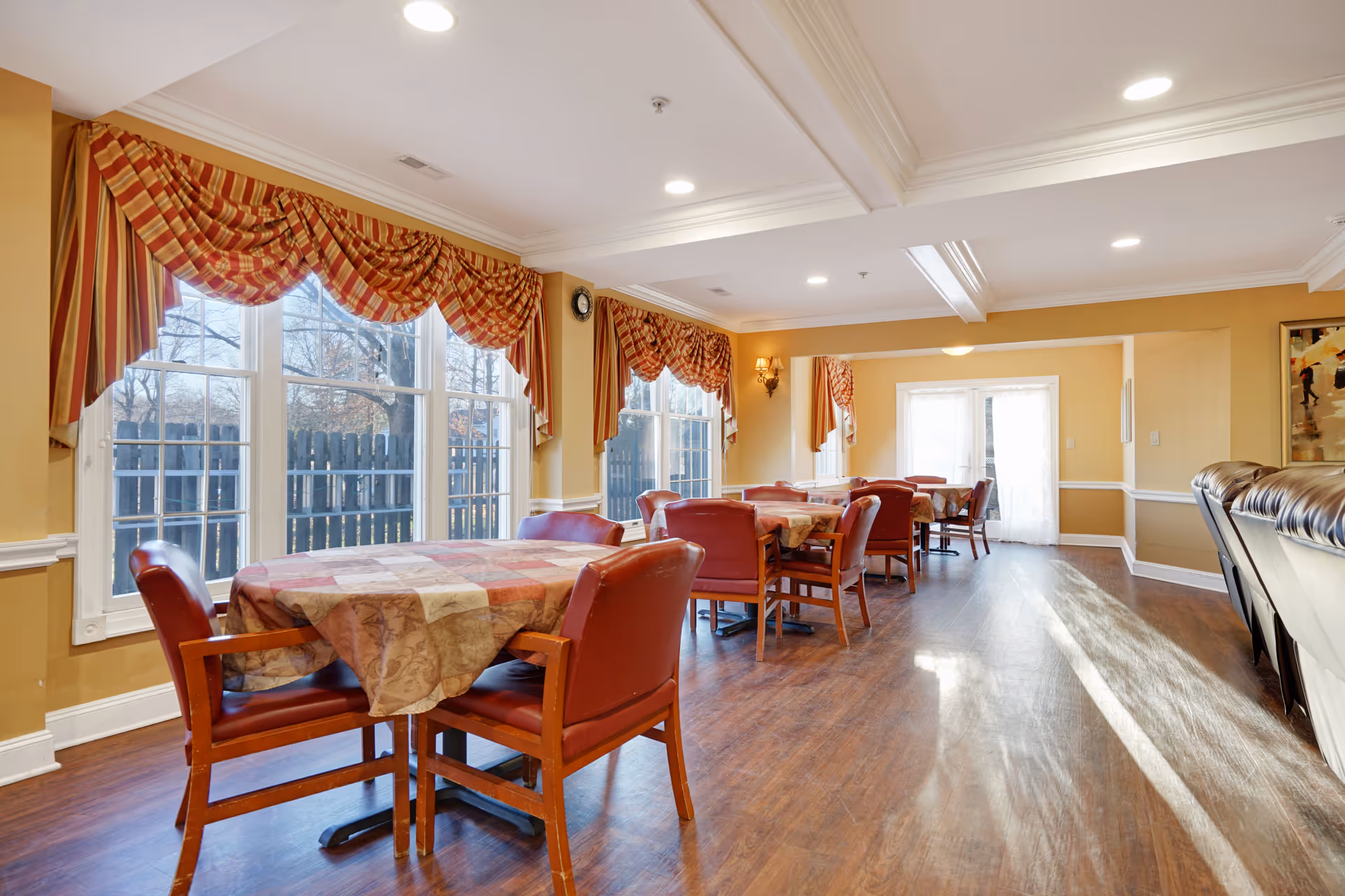 A bright dining room with several tables covered with patterned tablecloths and surrounded by red cushioned chairs. Large windows with red and gold striped valances let in natural light. The room has wooden flooring, beige walls with white trim, and recessed ceiling lights. There is a door at the far end of the room and a few wall sconces providing additional lighting.