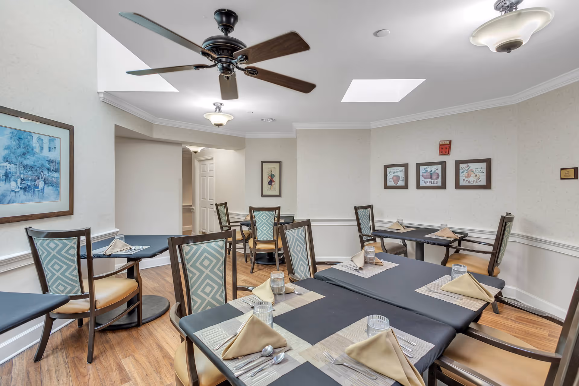 Small dining room with multiple set tables and chairs, ceiling fan, skylights, and framed wall art.