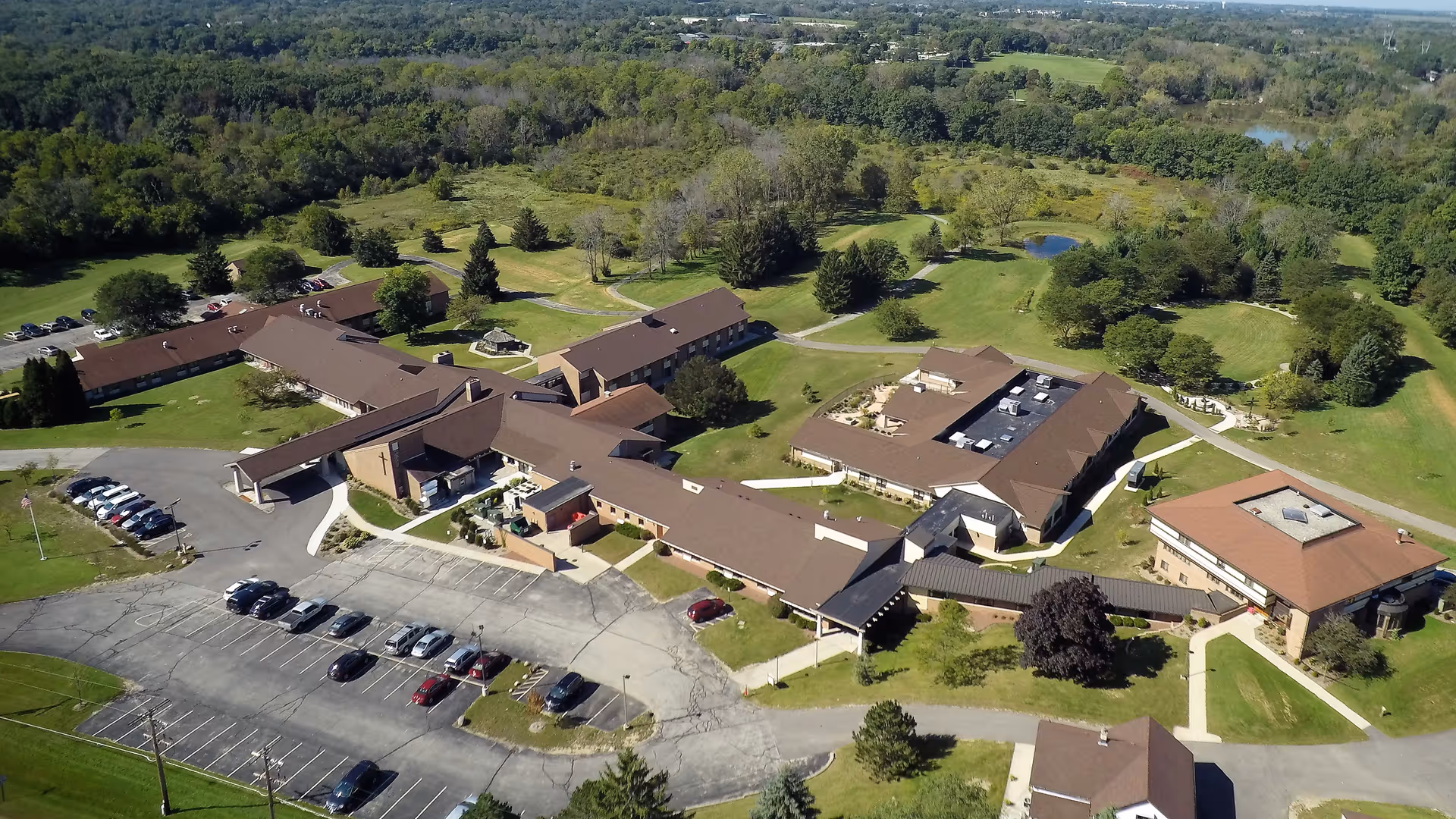 Aerial view of St Monica's Senior Living facility showing multiple connected buildings with brown roofs surrounded by green lawns, trees, and parking lots. The facility is set in a rural area with dense forest in the background.