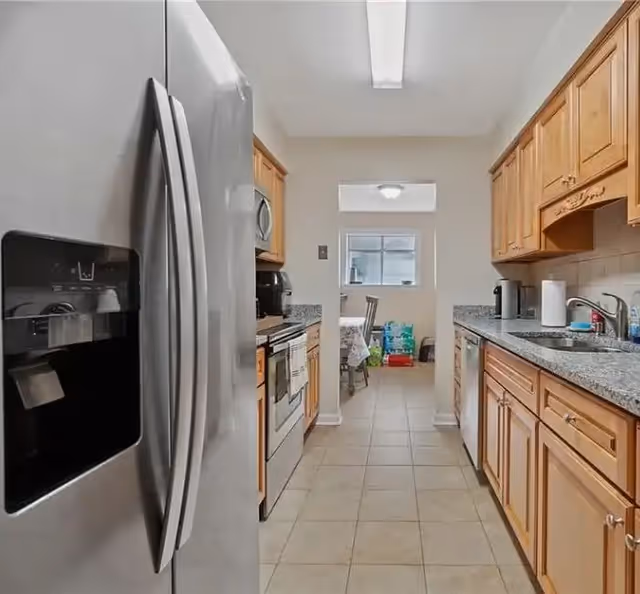 Galley-style kitchen with a stainless steel refrigerator, oven, granite countertops and wooden cabinets leading to a small dining area.