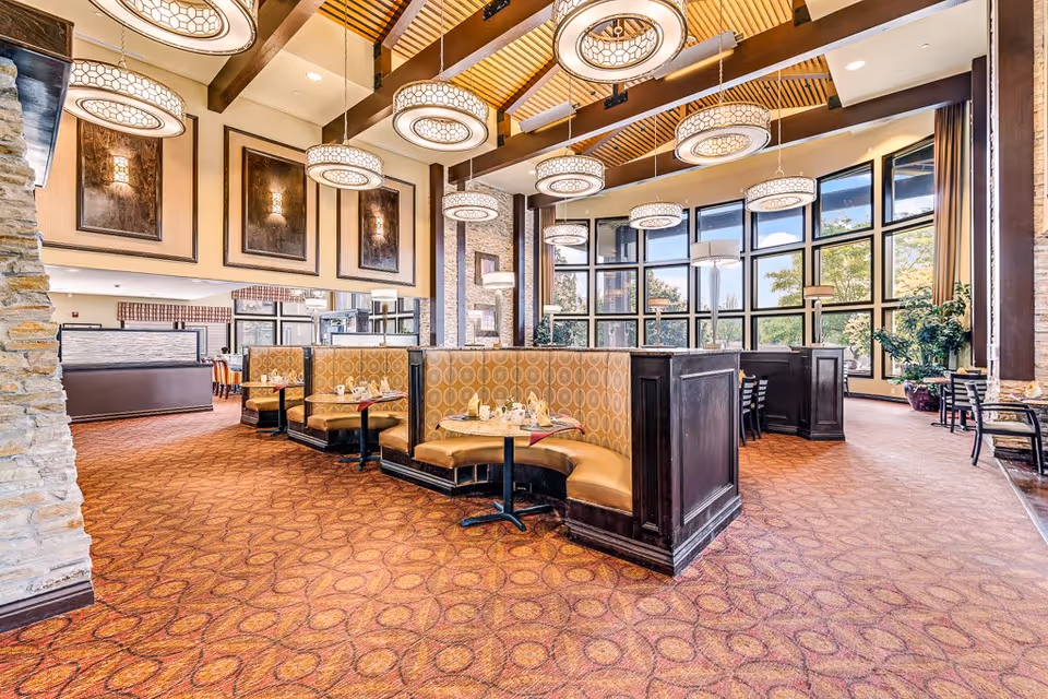 A spacious dining area with curved booth seating upholstered in patterned yellow fabric, round tables set with napkins and cups, large windows letting in natural light, and modern circular pendant lights hanging from a high ceiling with wooden beams.