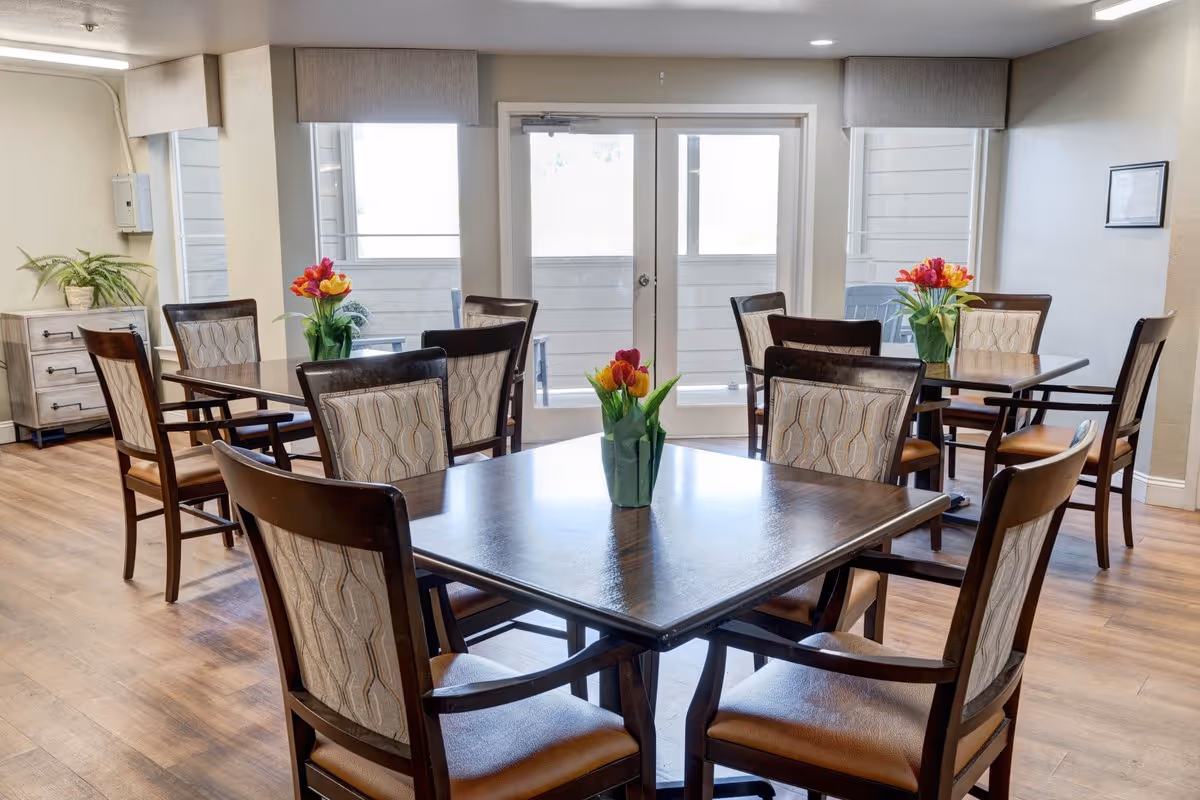 A bright dining room with wooden floors and multiple square tables, each surrounded by four cushioned chairs. Each table has a vase with colorful flowers. Large windows and glass doors let in natural light, and there is a small cabinet with a plant on the left side of the room.